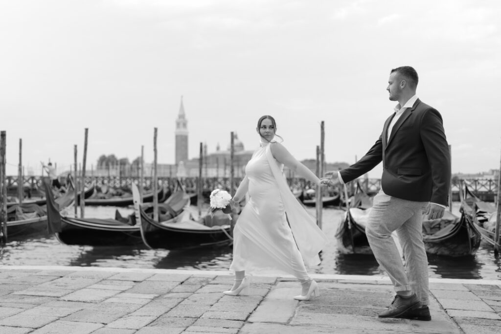 Black and white photo of a couple walking by gondolas during their Venice elopement
